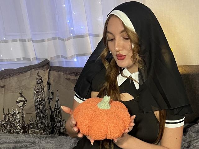 JoslynMensick with long brown hair in a nun outfit, holding an orange, fluffy pumpkin with a green stem, looking down with red lipstick, sitting on a bed.