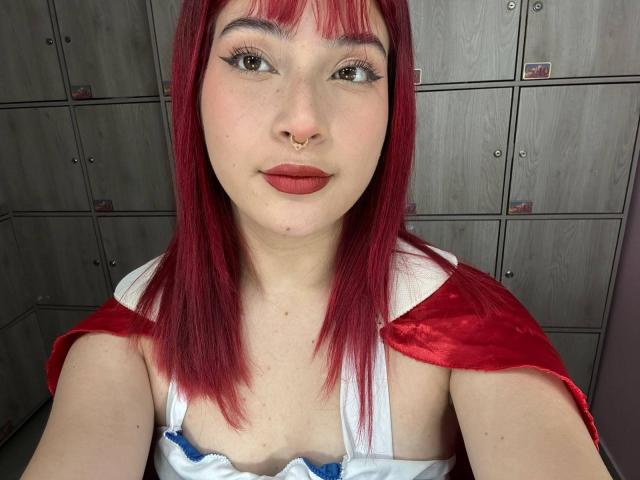 HaileyParck with red hair, nose ring, and red lipstick, wearing a white top with blue trim and red cape, showing her tits. She's smiling at the camera.