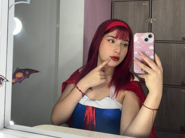 HaileyParck with red hair and headband, pouting with finger on lip, taking mirror selfie, wearing blue and red dress, showing pale skin and red lipstick.