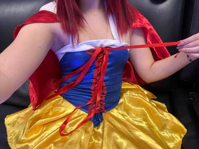 HaileyParck in a sexy Snow White cosplay, showing her shiny yellow skirt, tight blue corset with red laces, and red cape, holding a red ribbon with her hand.