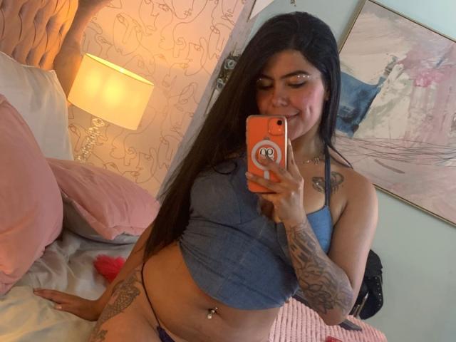 CatalinaReiyna, con cabello negro largo, vestida con un top corto azul, tanga morada y tatuajes, tomando una selfie frente al espejo, mostrando sus pechos y ombligo, en un dormitorio rosa..
