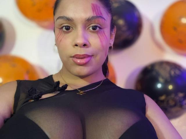 TeylorJenkins with dark hair, pink face marks, and black sheer top, showing large boobs, wearing a gold necklace, looking at camera.