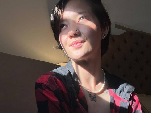 A LissyLuxe tem cabelo preto curto, um piercing no nariz, alargadores de orelha pretos, um colar de pérolas e uma camisa xadrez vermelha, sorrindo com o sol no rosto..