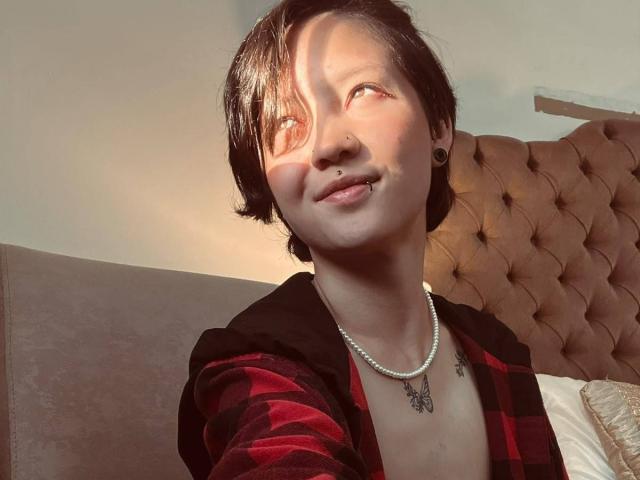 LissyLuxe, avec ses cheveux noirs courts, son anneau de nez et son piercing aux lèvres, porte une chemise à carreaux rouges, un collier de perles et des élargisseurs d'oreille noirs, et sourit au soleil. Un tatouage est visible sur le haut de sa poitrine..