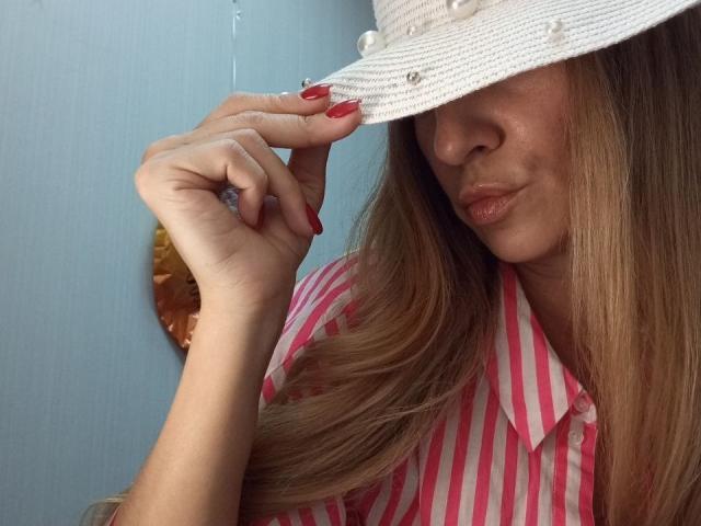 ViktoriiaLu, avec de longs cheveux bruns, des ongles rouges, portant une chemise rose à rayures et un chapeau blanc avec des perles, touchant le bord du chapeau, lèvres pincées, mur bleu en arrière-plan..