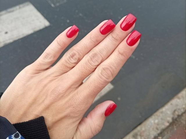 La mano di ViktoriiaLu, con lo smalto rosso lucido, le dita leggermente divaricate, che mette in mostra le unghie ben curate e lucide. La sua pelle è chiara e indossa una manica nera..