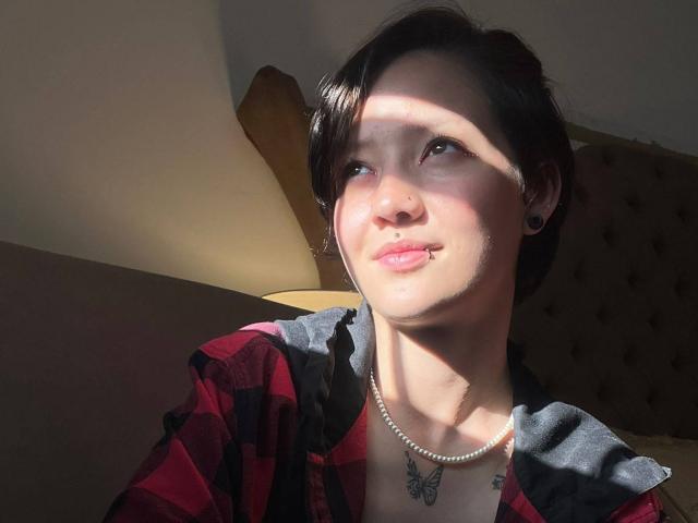 LissyLuxe with short black hair, red plaid shirt, black ear plugs, pearl necklace, butterfly tattoo, light shadow on face, slight smile, and sunlight highlighting her skin.