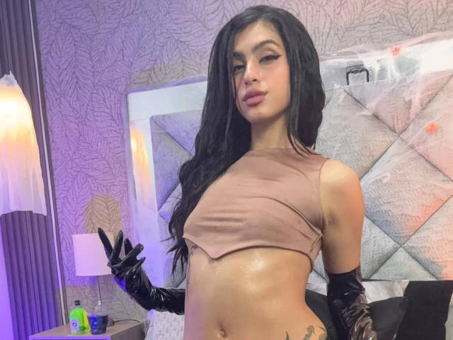 LilithDesire with long black hair, wearing a pink crop top, black gloves, and showing her bare tits and belly button. She's standing in a bedroom, looking at the camera.