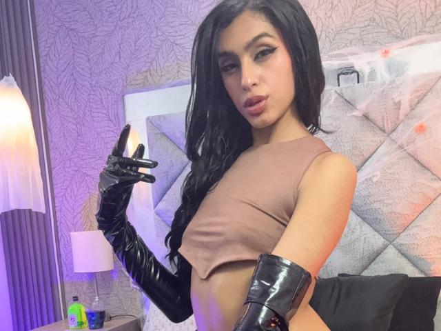 LilithDesire with long black hair, wearing a brown crop top, black latex gloves, and black latex boots, making a rock-and-roll hand gesture, showing her small tits.