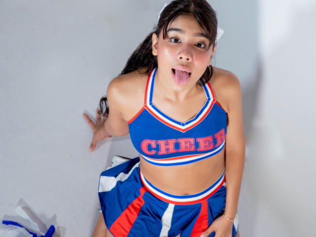 AmyLambert, with brown hair in pigtails, wearing a blue and red cheerleader outfit, sticking out her tongue, hands on white floor, midriff exposed, looking up at camera.