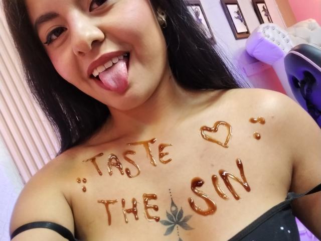 AlessiaVeega with long black hair, tongue out, caramel sauce on her chest, tattoo below her tits, wearing a black strap top, smiling at the camera.