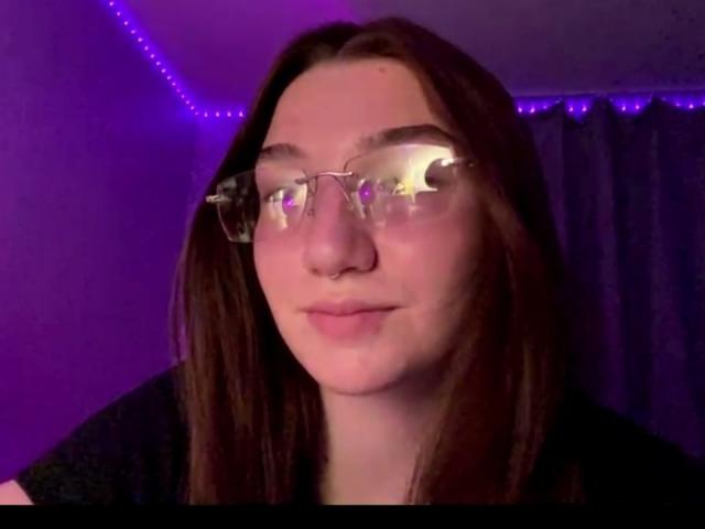 SunnyKlusman with clear glasses, reflecting purple lights, has straight brown hair, pale skin, and a slight smile. She's wearing a black shirt in a purple-lit room.