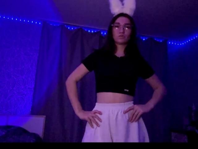 SunnyKlusman with white bunny ears, glasses, black crop top, and white skirt, standing with hands on hips, in a blue-lit room. Midriff exposed, looking serious.