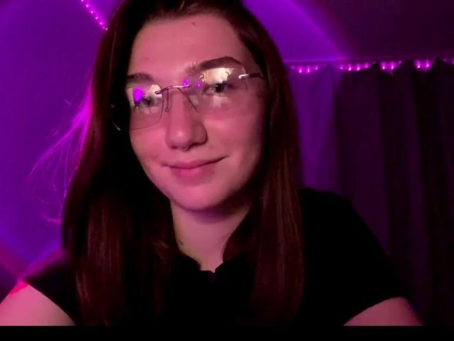 SunnyKlusman with glasses, smiling slightly, brown hair, wearing a black shirt, purple-lit background, reflection on glasses, soft smile, medium skin, subtle makeup, gentle expression.