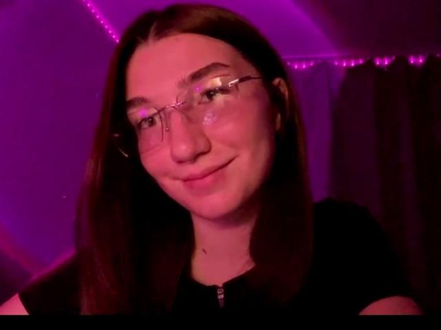 SunnyKlusman with long brown hair, glasses, smiling, pinkish-purple light, wearing black shirt, soft smile, fair skin, slight blush on cheeks, subtle makeup, relaxed vibe.
