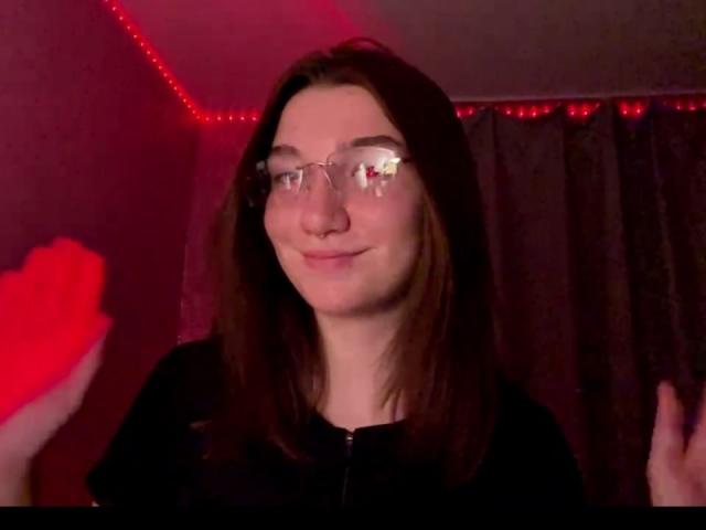 SunnyKlusman with glasses, brown hair, black shirt, holding a red heart, smiling slightly, red LED strip lights in background, right hand raised.