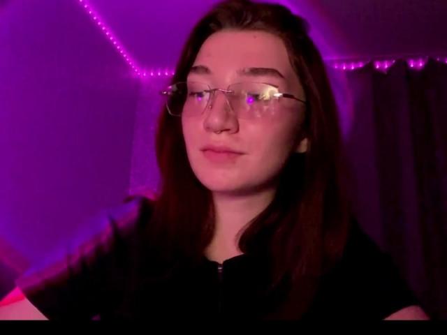 SunnyKlusman with red hair, wearing glasses, black shirt, and a slight smile, under purple neon light, looking straight at the camera. Her tits are partially visible.