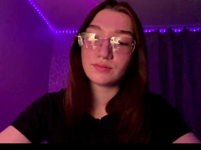 SunnyKlusman has pale skin, brown hair, clear glasses, and a black shirt. She's looking slightly to the side with a neutral expression in purple-lit room.