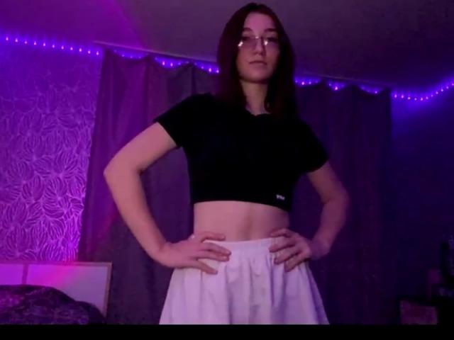 SunnyKlusman with hands on hips, wearing glasses, black crop top, white pants, standing in dimly lit room with purple LED strip lights, mid-length brown hair, confident expression.