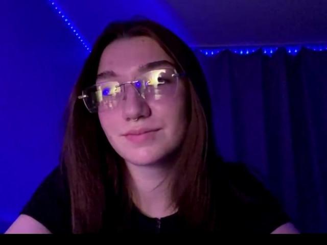 SunnyKlusman with glasses, brown hair, nose ring, and slight smile, wearing a black shirt, in a blue-lit room. Her face is centered, looking slightly to the right.