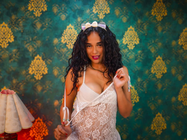 ValeryNicolls with curly black hair, wearing white lace lingerie, holding straps, smiling, pink lipstick, and a white bow headpiece, standing in front of a green and gold patterned wall.