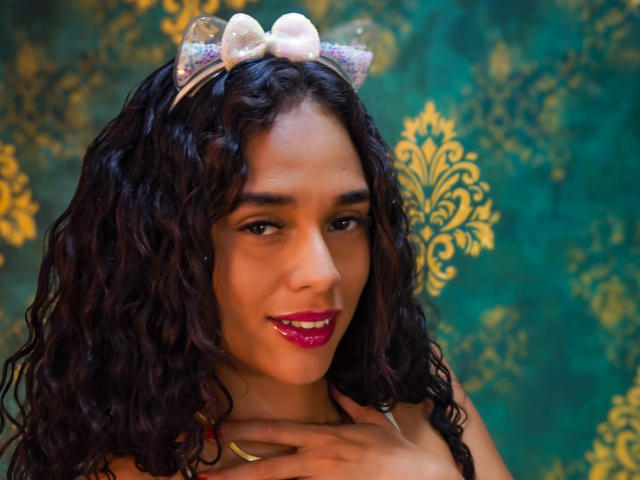 ValeryNicolls has curly dark hair, a white bow with sparkles, pink lipstick, and a hand near her neck. She's smiling with brown eyes and a medium skin tone.