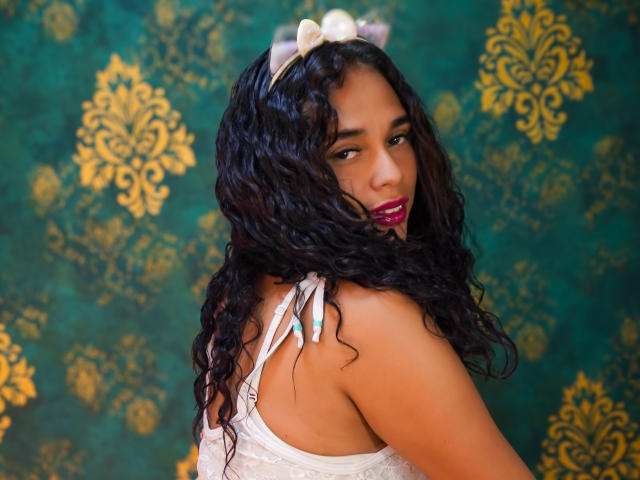 ValeryNicolls with curly black hair, white lace dress, and pink lipstick, looking over shoulder, showing her tits, green-blue eyes, and a cute smile, with a gold patterned background.