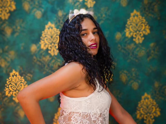 ValeryNicolls with curly black hair, wearing a white lace top, hands on hips, gold headpiece, and pink lipstick, looking over shoulder, displaying small, perky tits.