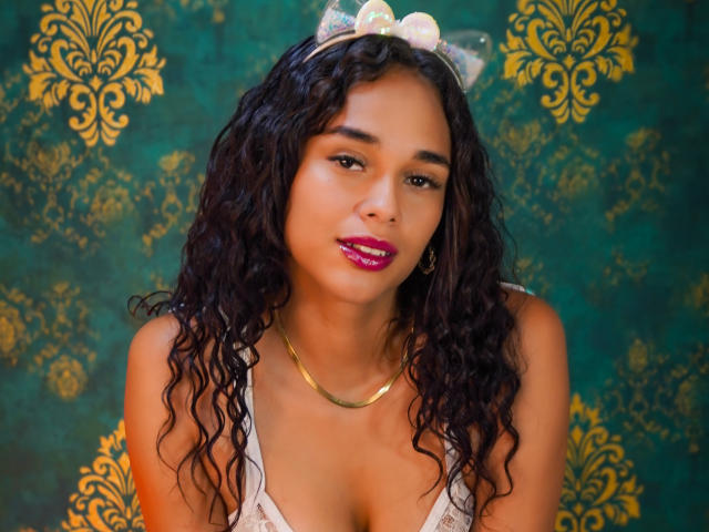 ValeryNicolls has curly black hair, pink lipstick, gold necklace, and white bow headband, showing cleavage in a lace top, with a sexy, confident expression.
