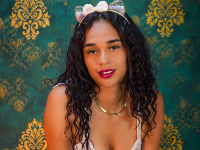 ValeryNicolls has curly black hair, wearing a white bow headband, gold necklace, and red lipstick, with her tits slightly visible in a white top, smiling at the camera.
