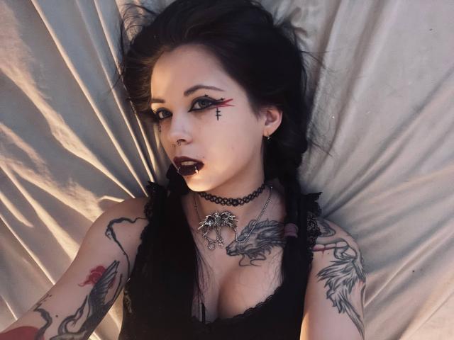 BellaFavian with dark makeup, black lipstick, and red eyeliner, lying on a bed. She has tattoos, a black choker, and a dragon necklace, wearing a black lace top.