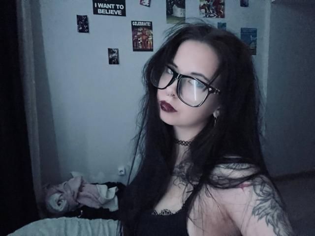BellaFavian with long black hair, glasses, dark lipstick, choker, tattoos, black top, showing tits, looking at camera, in dimly lit room, messy bed in background.
