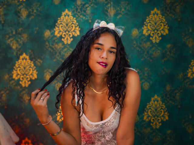 ValeryNicolls with curly black hair, wearing a white lace dress, holding a lock of hair, gold necklace, pink lipstick, green patterned wallpaper, and a white cat ear headband.