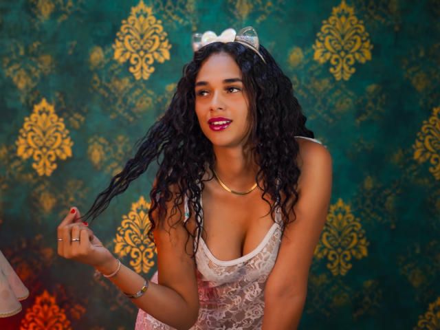 ValeryNicolls with curly black hair, wearing a white lace top, gold necklace, and silver tiara, holding her hair, smiling with pink lipstick, showing her tits, against a green patterned background.