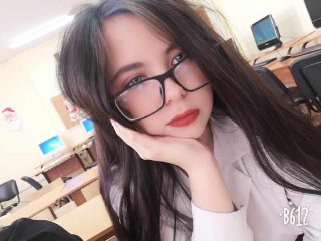BellaFavian with long black hair, wearing glasses and a white shirt, resting her head on her hand, showing her red lips and soft skin, in a bright office.