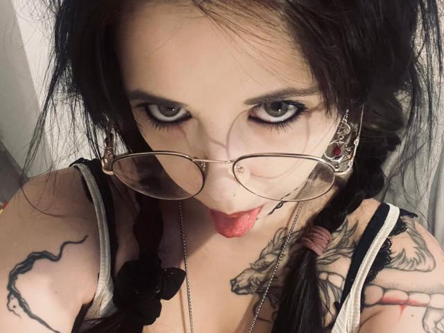 BellaFavian with black hair in braids, wearing glasses, sticking out her tongue, tattoos on arms, and a black strap, looking up with blue eyes. She's got a sexy look.