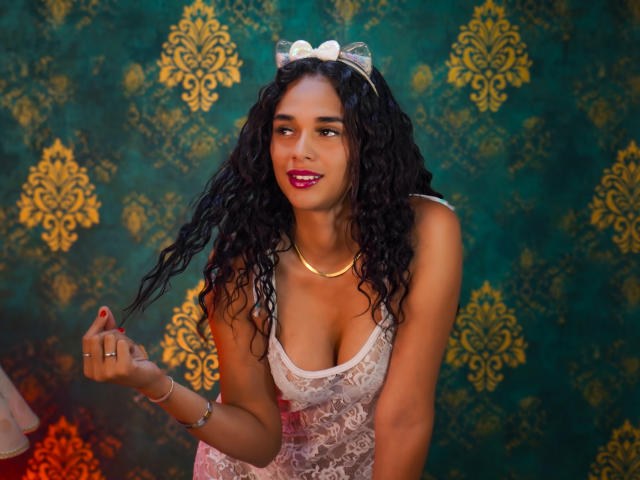 ValeryNicolls has curly black hair, pink lipstick, and a white lace dress with a bow. She's holding her hair, showing her small tits, and wearing a gold necklace.