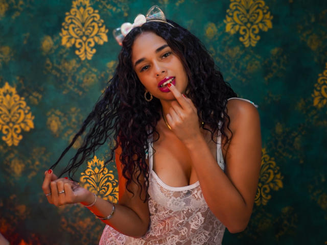 ValeryNicolls with curly black hair, wearing a white lace top, finger in mouth, gold hoop earrings, ring, and white headband, holding hair, showing cleavage, smiling, dark skin.