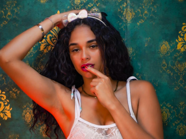 ValeryNicolls with curly black hair, white lace top, and a pink bow headband, finger on red lips, arm raised, green and gold patterned background, red light on side.