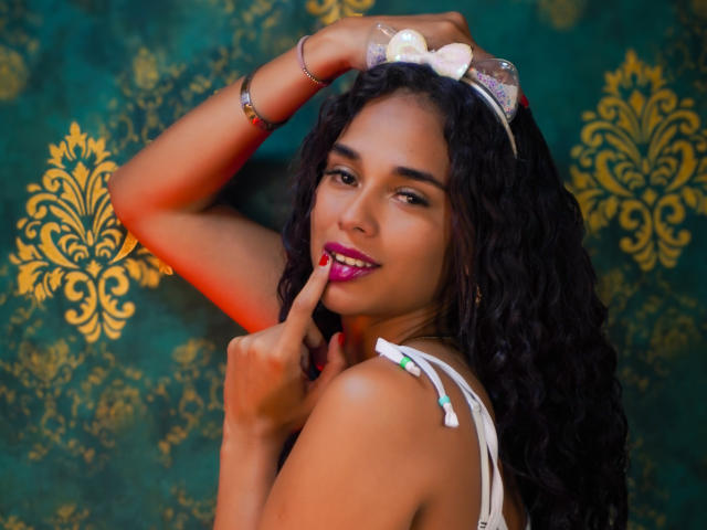 ValeryNicolls with curly black hair, pink lipstick, finger touching lips, white strap dress, silver bracelet, headband with white bow, green and gold patterned background, smiling, sexy.