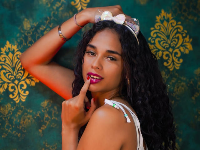 ValeryNicolls with curly black hair, wearing a white bow headband and white top, has a finger to her pink lipstick, looking sexy with a green and gold patterned background.