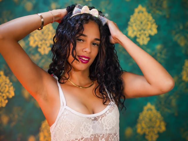 ValeryNicolls with curly black hair, wearing a white lace top, gold necklace, and bracelet, hands in hair, looking at camera, purple lipstick, green and gold patterned background.
