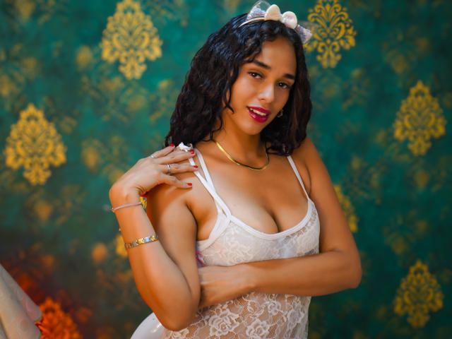 ValeryNicolls with curly black hair, wearing a white lace top, showing her tits, pink lipstick, gold necklace, and cat ears, smiling, with green and gold patterned background.