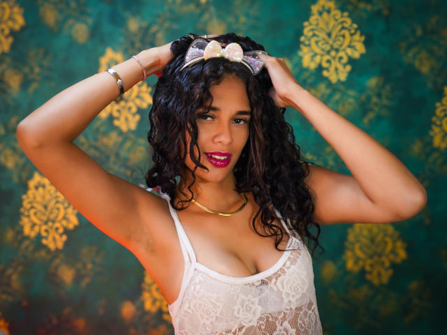 ValeryNicolls with curly black hair, wearing a white lace top, gold necklace, and pink lipstick, hands in hair, looking at camera, showing her tits. Green and gold background.