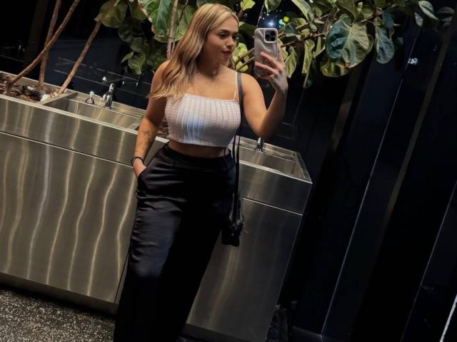 ZoeVibe met blond haar, een witte top zonder bandjes, zwarte broek, handen in de zakken, die een selfie maakt in een donkere badkamer, een telefoon vasthoudend, een kleine camerastrap, een groene plant op de achtergrond..