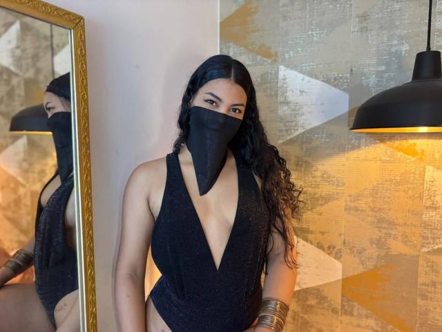 Lalila, una ragazza moro di capelli lunghi neri, indossa un costume intero nero a V profondo, una maschera nera che le copre naso e bocca, braccialetti dorati e, sullo sfondo, una lampada a sospensione nera..