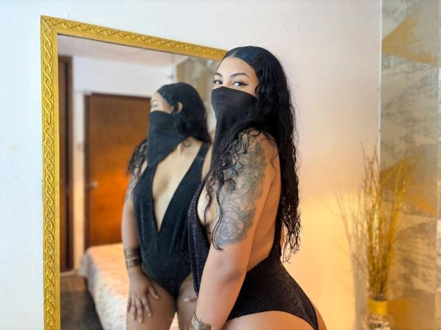 LalilaBrunette, avec de longs cheveux noirs, portant un masque noir et de la lingerie noire à décolleté plongeant, montrant son bras tatoué et ses courbes dans un miroir, ses seins étant partiellement visibles..