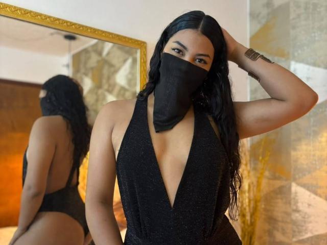 LalilaBrunette with long black hair, wearing a deep black dress with a plunging neckline, black mask, and gold bracelet, hand in hair, reflected in a gold-framed mirror.