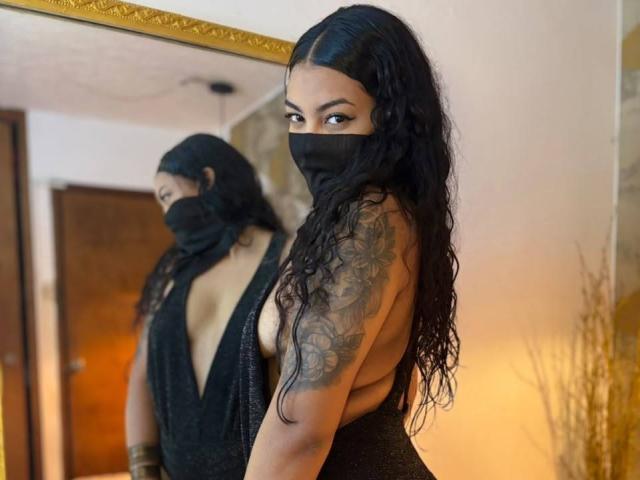 Lalila Brunette, avec de longs cheveux noirs ondulés, portant un masque noir et une robe noire à décolleté en V profond, montrant un tatouage sur son bras droit, se tient devant un miroir..