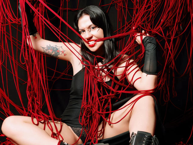 GoddessLuxliv, com cabelo preto, batom vermelho e tatuagem, vestindo um vestido preto e luvas, mordiscando de forma divertida uma corda vermelha, com os seios e a vulva levemente visíveis, cercada por cordas..